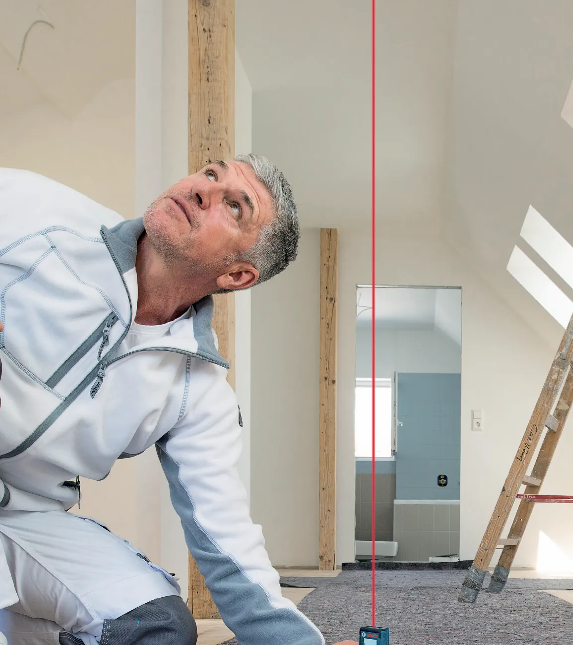 Handwerker misst im ausgebauten Dachgeschoss mit einem Laser-Entfernungsmesser die Raumhöhe, eine rote Laserlinie führt zur Decke