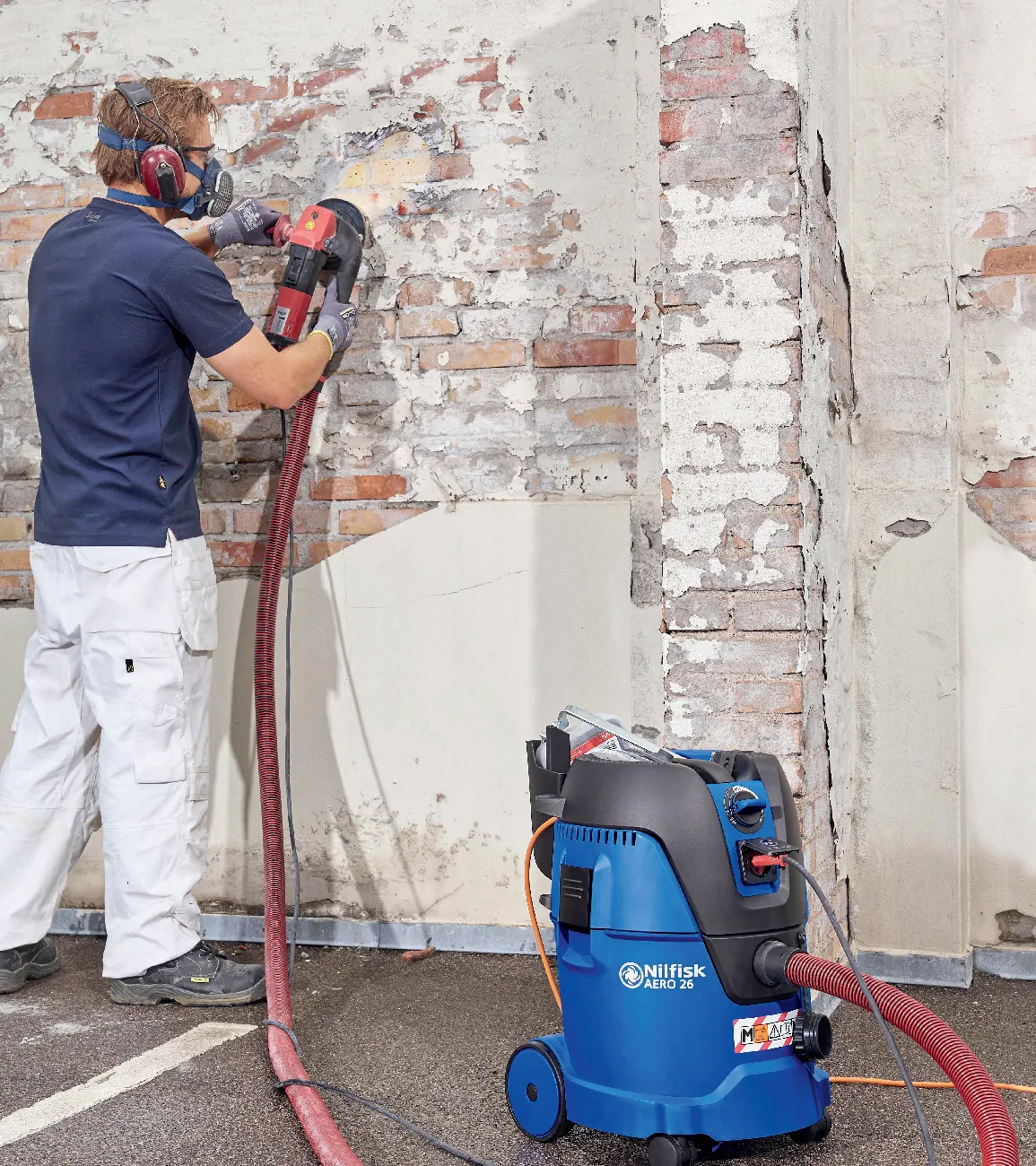 Handwerker mit Gehörschutz und Atemschutzmaske schleift eine Wand ab, das Gerät ist über einen Schlauch an einen Sicherheitssauger angeschlossen