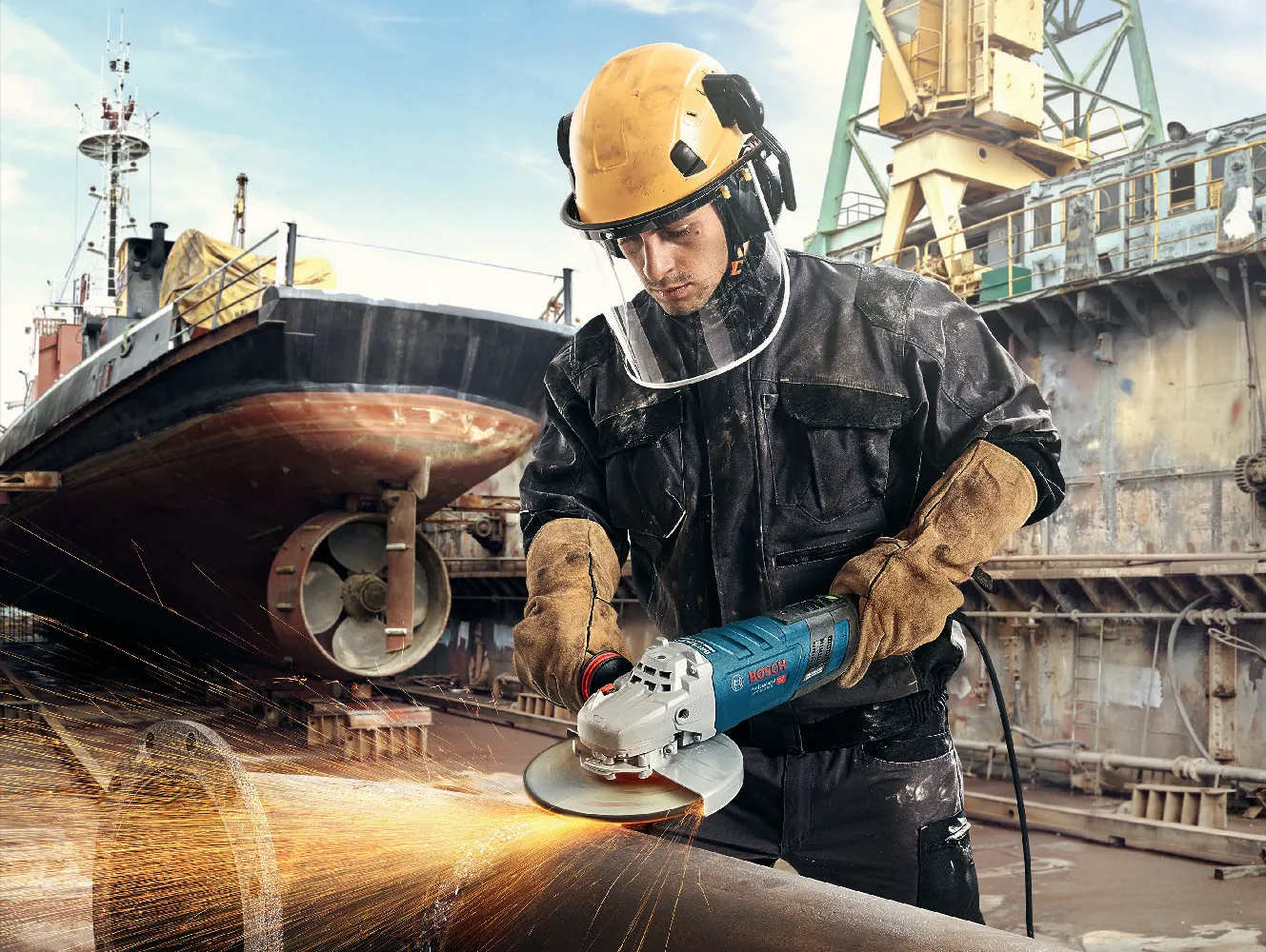Arbeiter mit Schutzhelm, Gesichtsschild und Gehörschutz schleift mit einem großen Winkelschleifer an einem Stahlrohr auf einer Werft